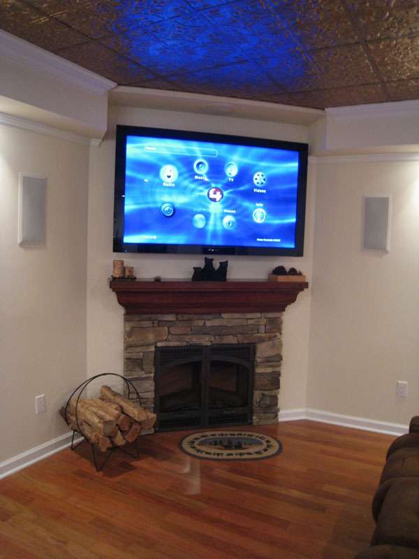 Corner TV over a stone fireplace showing a home control menu, with speakers set into the walls