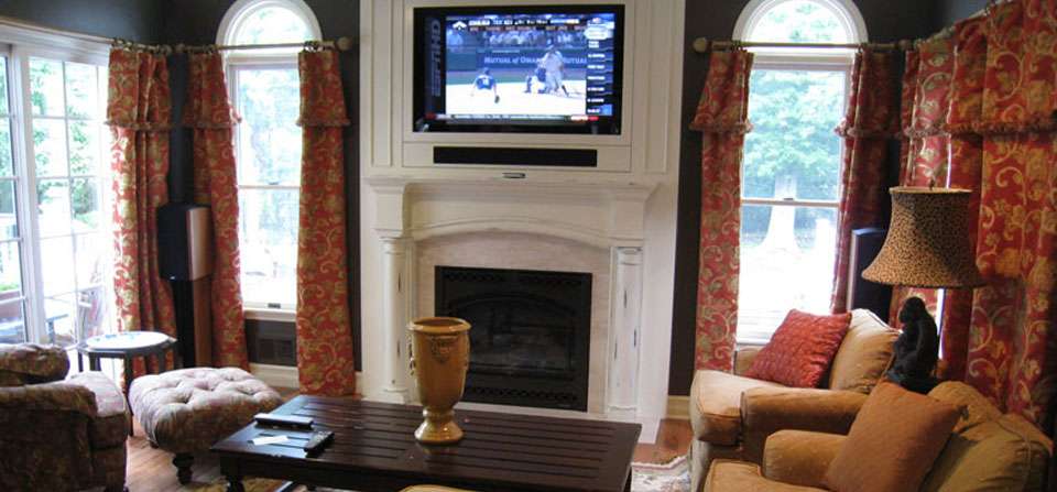 Family room with a TV above a white fireplace, tall windows and drapes on both sides